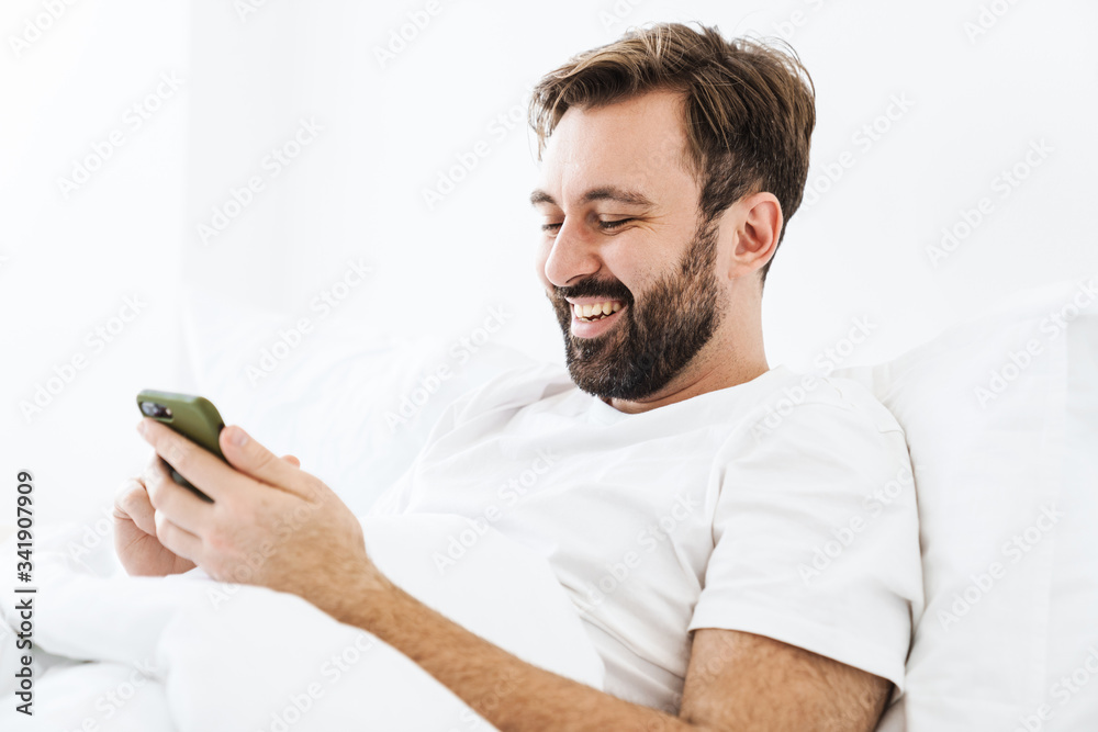 Image of young unshaven caucasian man smiling and using cellphone while lying in bed at home Image of young unshaven caucasian man smiling and using cellphone while lying in bed at home