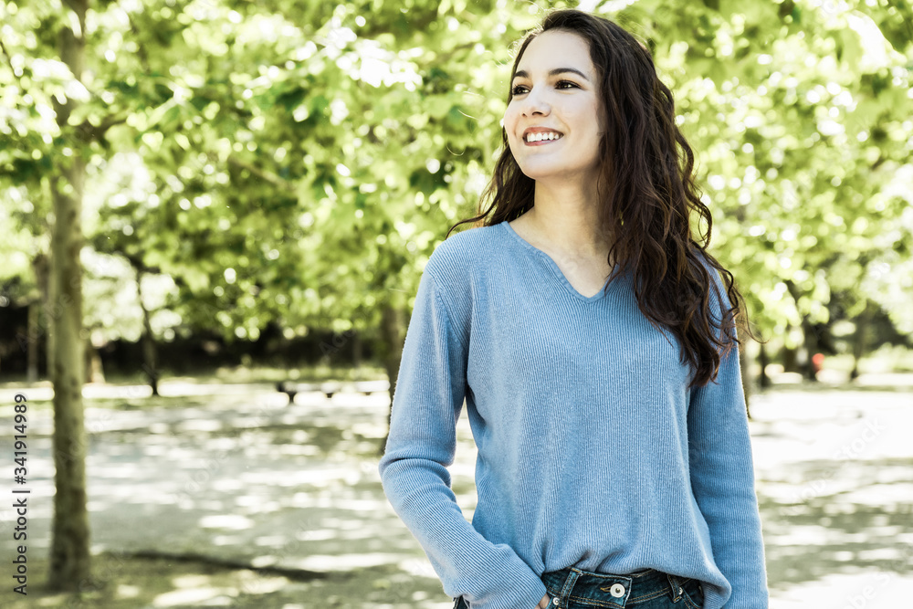 Smiling young woman standing in spring park and looking aside. Attractive young lady with long hair holding hand in pocket while posing in spring park. Female beauty concept Smiling young woman standing in spring park and looking aside. Attractive young lady with long hair holding hand in pocket while posing in spring park. Female beauty concept