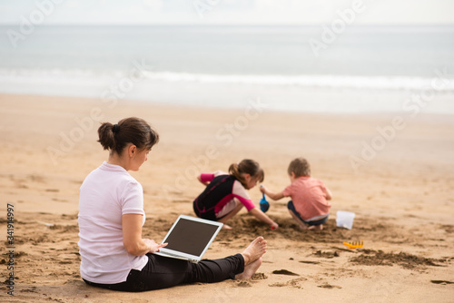 Busy mom teleworking on laptop while children playing beside shore © Martinan
