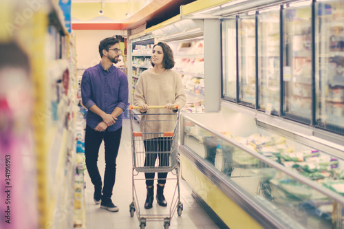 Couple walking and talking in grocery store. Happy young man and woman with shopping trolley choosing products in supermarket. Shopping concept