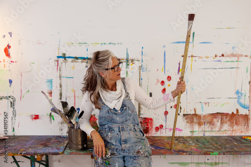 proud older artist woman, in her fifties with grey hair and black glasses holds a big paintbrush