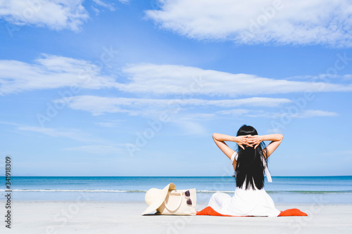 Asian woman, long black hair, wore white dress and hat sitting on the beach near the woven bags and sunglasses and facing back by the sea with copy space blue sky,summer holiday and vacation concept © Ann Patchanan