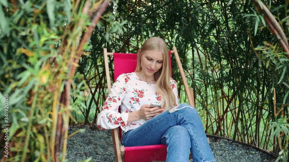 Young woman using smartphone in plant gazebo. Beautiful young lady smiling and typing message on smartphone while sitting inside amazing plant gazebo in park. Young woman using smartphone in plant gazebo. Beautiful young lady smiling and typing message on smartphone while sitting inside amazing plant gazebo in park.