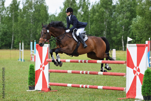 Young equestrian sports girl jumping obstacle with bay horse