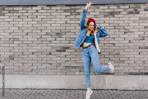 Full-length photo of enchanting lady in sport shoes and denim jacket dancing near brick wall. Outdoor shot of caucasian girl jumping on the street with happy face expression.