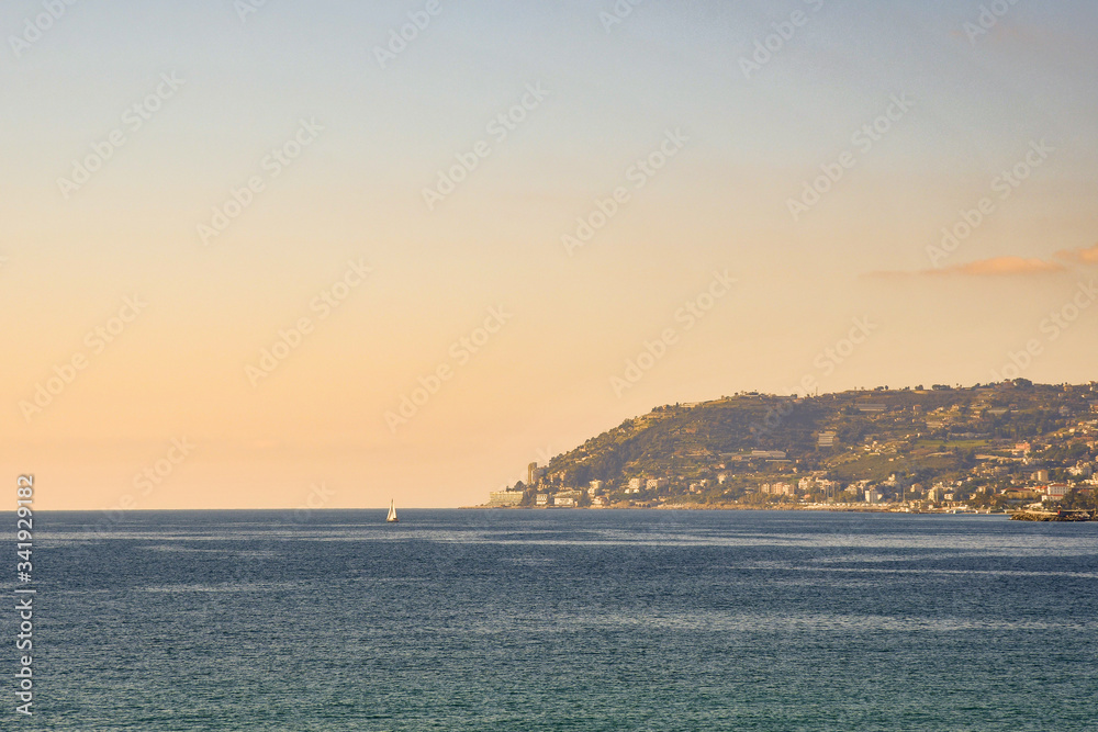 Fototapeta premium Mediterranean seascape with a cape and a sailboat on the sea horizon at sunset, Sanremo, Capo Nero, Imperia, Liguria, Italy