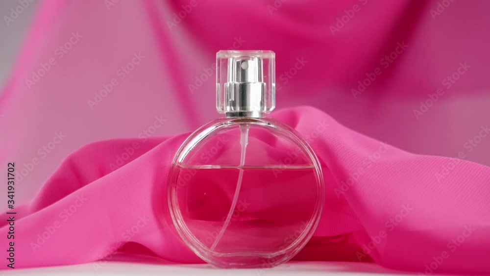 Oval surfaced perfume bottle with pink perfumes or essential oils is on the table. Pink fabric flutters around and waves in the air around the perfume bottle. Concept of aroma and smell. Close up Oval surfaced perfume bottle with pink perfumes or essential oils is on the table. Pink fabric flutters around and waves in the air around the perfume bottle. Concept of aroma and smell. Close up