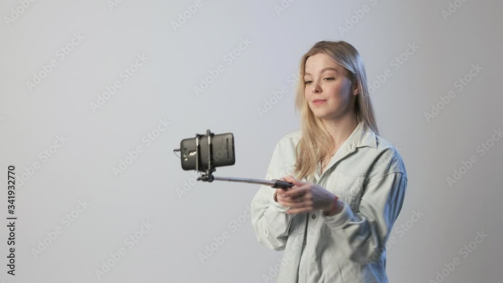 cute female records video by smartphone attached to selfie stick. blond girl filming vlog speaking, smiling, chatting with followers, using cellphone on white background. Beauty blogger recording blog