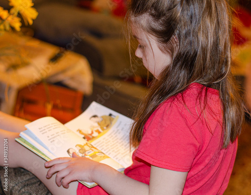 a girl child of 6 years old is sitting on the sofa and reading a book at home. Home schooling, self-study.