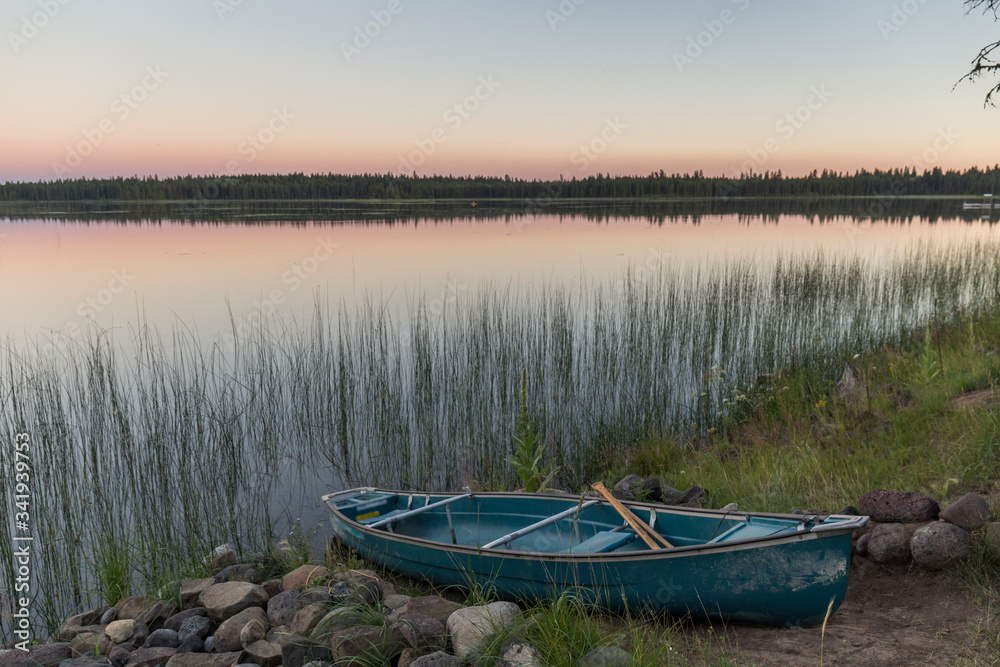 wooden boat stranded next to a calm water lake in which the orange colors of the sunset and the trees in the background are reflected, next to which reeds grow wooden boat stranded next to a calm water lake in which the orange colors of the sunset and the trees in the background are reflected, next to which reeds grow