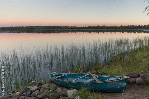 wooden boat stranded next to a calm water lake in which the orange colors of the sunset and the trees in the background are reflected, next to which reeds grow