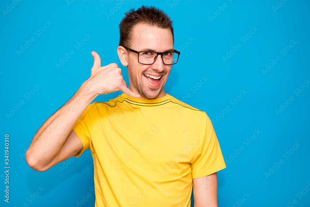 Good looking young man in glasses and yellow shirt gesturing looking at camera while standing against blue background Good looking young man in glasses and yellow shirt gesturing looking at camera while standing against blue background