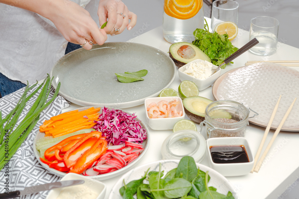 The process of cooking spring rolls - woman's hands putting basil on rice paper. Table with spring rolls ingredients and water with oranges. The process of cooking spring rolls - woman's hands putting basil on rice paper. Table with spring rolls ingredients and water with oranges.