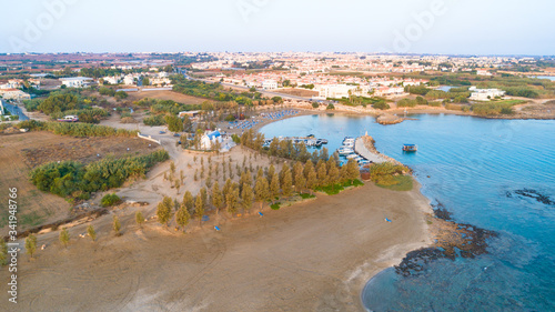 Aerial bird's eye view of coastline sunset, landmark white washed chapel Agia Triada beach, Protaras, Famagusta, Cyprus from above. Ayia Trias bay church and a boat out of the small port at sunrise.