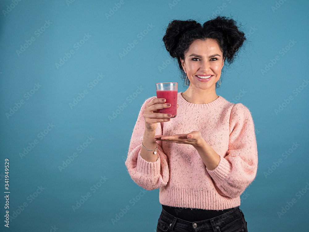Portrait of young woman with black curly hair in casual dress presenting a red smoothie isolated on blue background Portrait of young woman with black curly hair in casual dress presenting a red smoothie isolated on blue background