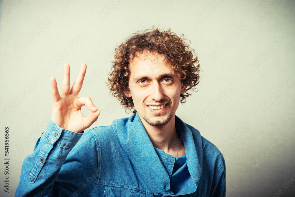 Curly young man makes a gesture okay. On a gray background Curly young man makes a gesture okay. On a gray background