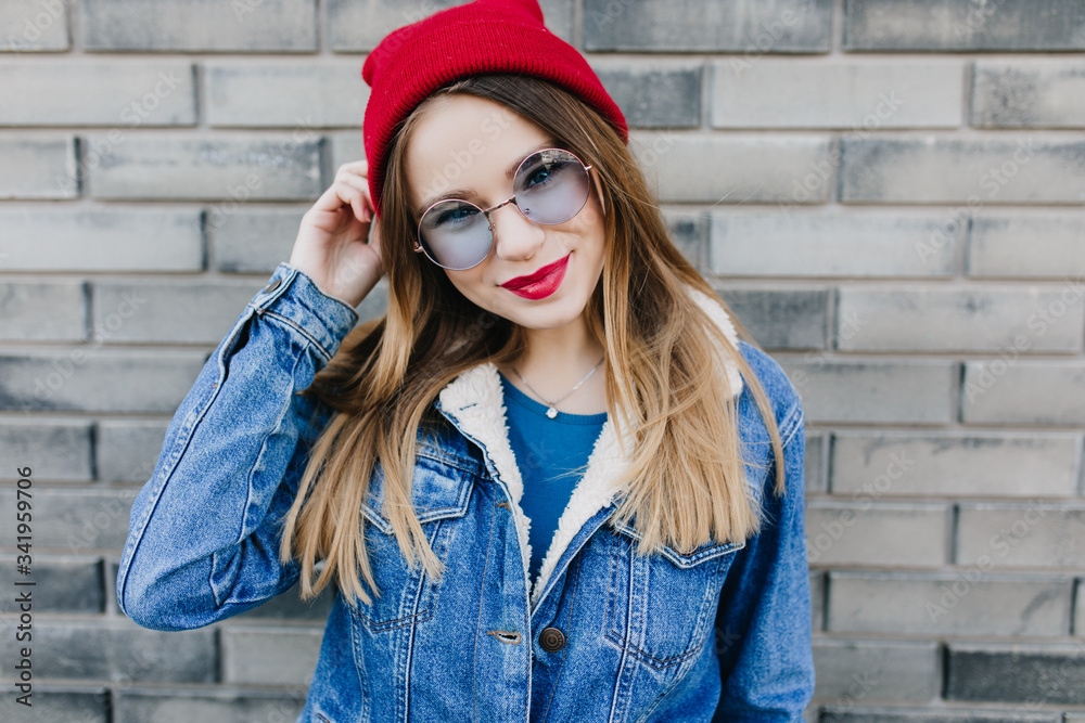White girl posing with sly smile on brick background. Ecstatic blonde woman in denim jacket touching her red hat and laughing. White girl posing with sly smile on brick background. Ecstatic blonde woman in denim jacket touching her red hat and laughing.