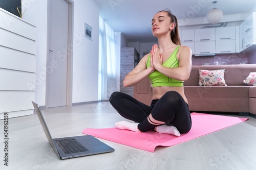 Young fitness woman in sportswear doing yoga exercises on mat in living room. Online workout, practicing and meditation at home