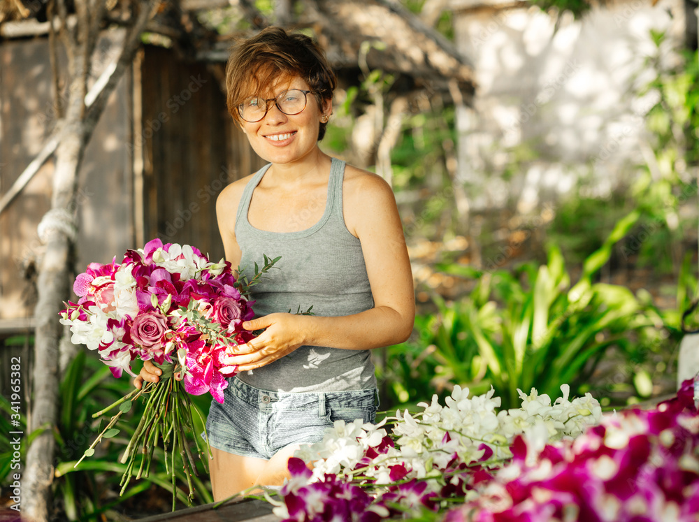 Beautiful young woman florist on the background of tropical greenery holds a bouquet of purple orchids.