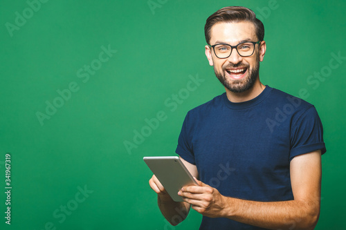 Happy young man in casual shirt and glasses standing and using tablet over green background