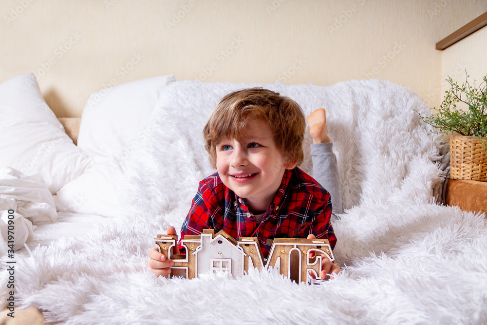 Obraz premium Happy little curly kid boy with smiley face lying on the bed at home. Stay at home quarantine coronavirus COVID-19 pandemic prevention. Wooden house inscription home.