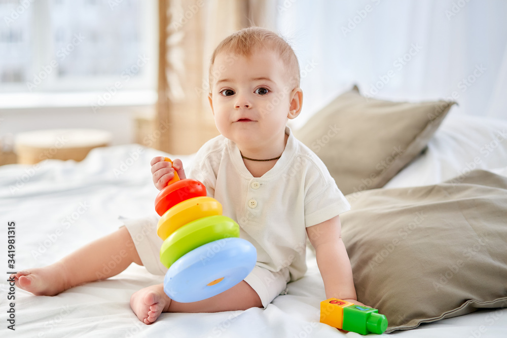 A charming baby plays with a peramid toy on a bed in a bright room. A charming baby plays with a peramid toy on a bed in a bright room.