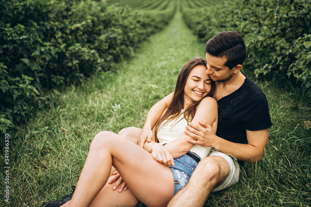 Woman with long hair and man sitting between currant bushes, hugging and kissing. Background of green currant plantations Woman with long hair and man sitting between currant bushes, hugging and kissing. Background of green currant plantations