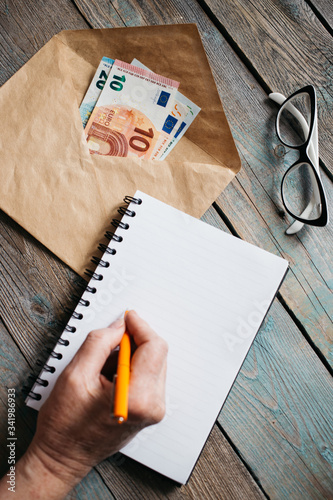 Close up of woman's hands writing in spiral notepad on wooden desktop, Euro banknotes in an envelope