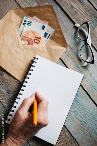Close up of woman's hands writing in spiral notepad on wooden desktop, Euro banknotes in an envelope