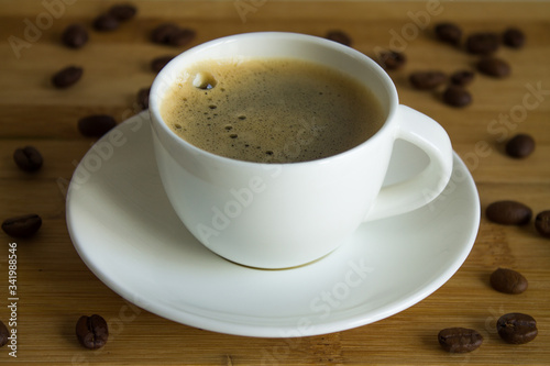 A small white Cup of fresh espresso with foam and a saucer close up on a brown wooden background