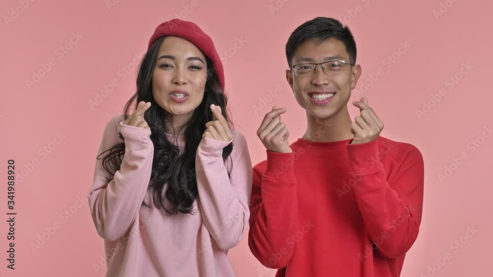 Young happy cute loving couple isolated over pink wall background make heart gesture.