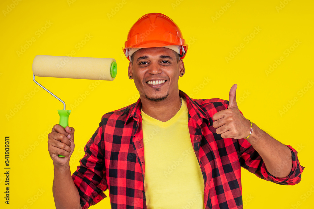 latin brazilian cleaning man wearing hardhat and painting the walls in studio yellow background