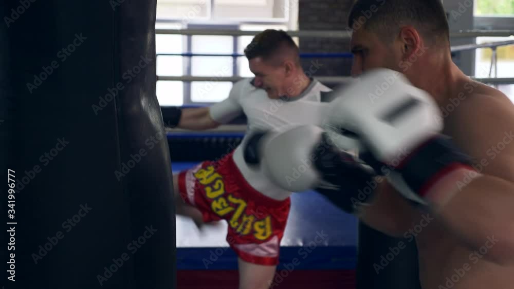 Two Professional Boxers Fiercely Punches and Kicks Punching Bags Near Boxing ring at Gym. Muay Thai Mixed Martial Arts Fighters Practicing Workout. 2x Slow motion 1/2 speed 60 fps 4K Two Professional Boxers Fiercely Punches and Kicks Punching Bags Near Boxing ring at Gym. Muay Thai Mixed Martial Arts Fighters Practicing Workout. 2x Slow motion 1/2 speed 60 fps 4K