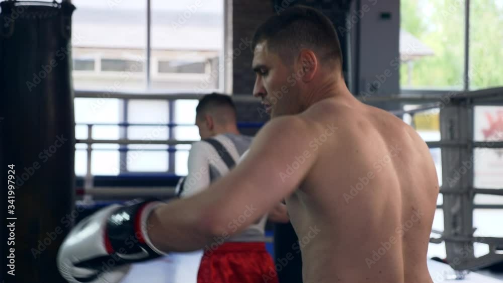 Two Professional Boxers Fiercely Punches and Kicks Punching Bags Near Boxing ring at Gym. Muay Thai Mixed Martial Arts Fighters Practicing Workout. 2x Slow motion 1/2 speed 60 fps 4K Two Professional Boxers Fiercely Punches and Kicks Punching Bags Near Boxing ring at Gym. Muay Thai Mixed Martial Arts Fighters Practicing Workout. 2x Slow motion 1/2 speed 60 fps 4K