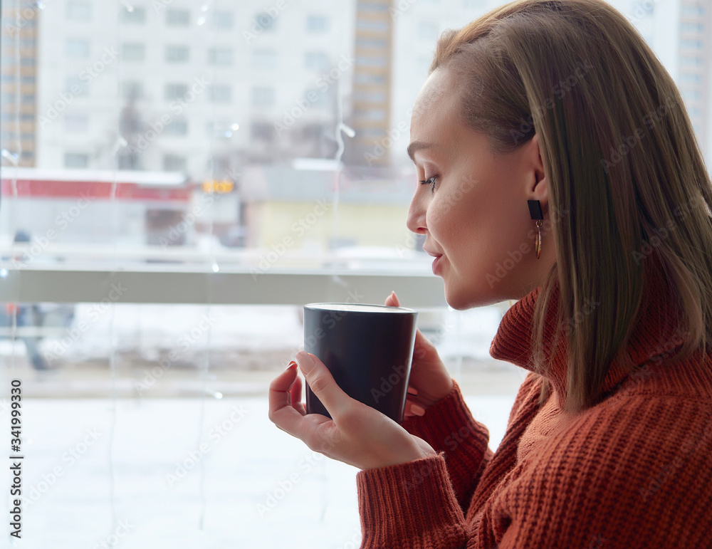 a dark Cup of coffee in the hands of a girl in a brown sweater. large window in the background. close up.