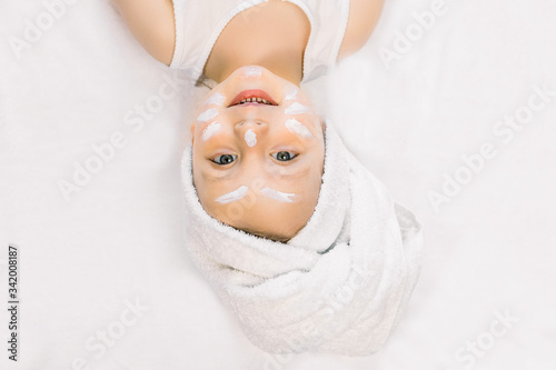Cute little girl with hair wrapped in bath towel, lying on white bed with cream on her face after bath or shower. Looking at the camera and smiling. Facial spa procedures, baby hygiene