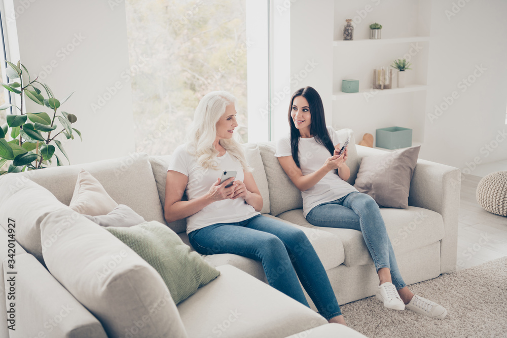 Portrait of two nice attractive lovely cheerful cheery women sitting on divan using digital device web app spending free time day weekend in white light interior house flat apartment Portrait of two nice attractive lovely cheerful cheery women sitting on divan using digital device web app spending free time day weekend in white light interior house flat apartment
