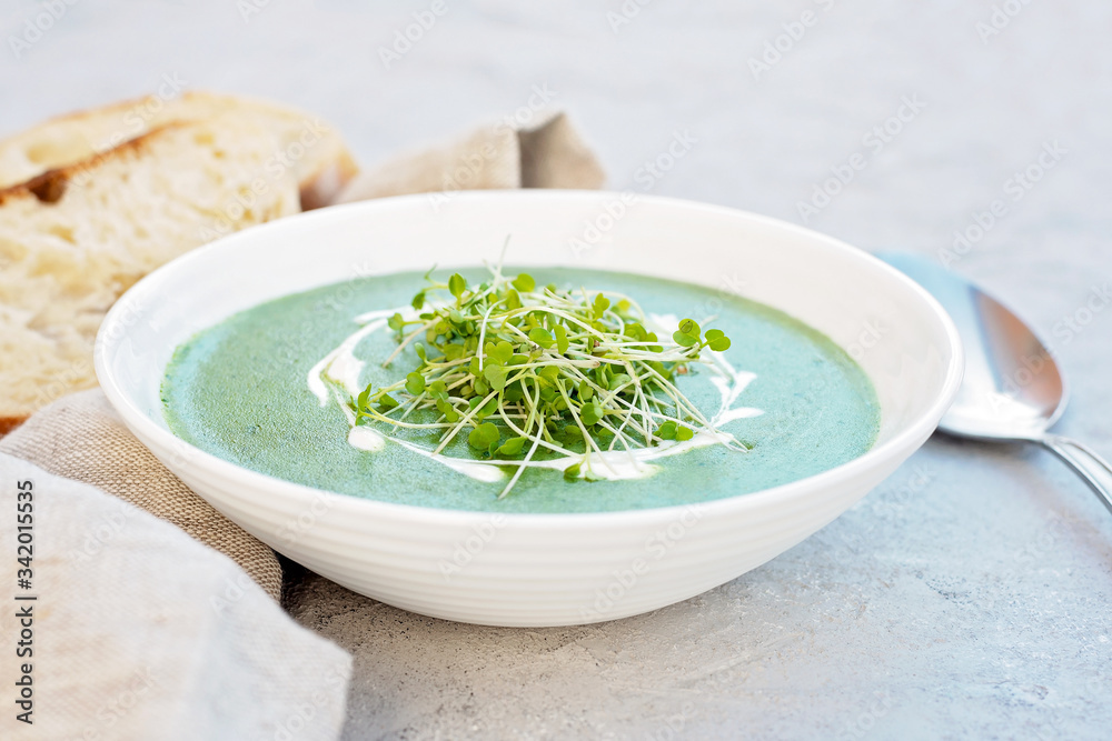 Cream soup of fresh spinach with cream and arugula microgreen in a white plate with ciabatta bread on a light gray background. Healthy and diet food. Cream soup of fresh spinach with cream and arugula microgreen in a white plate with ciabatta bread on a light gray background. Healthy and diet food.