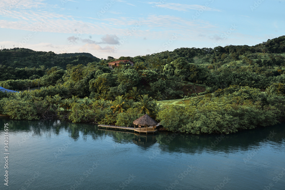 Tropical island view from the deck of cruise ship. Roatan island in Honduras with green trees in jungle woods and small wooden hut and pier. Travel landscape background. Tropical adventure. Tropical island view from the deck of cruise ship. Roatan island in Honduras with green trees in jungle woods and small wooden hut and pier. Travel landscape background. Tropical adventure.
