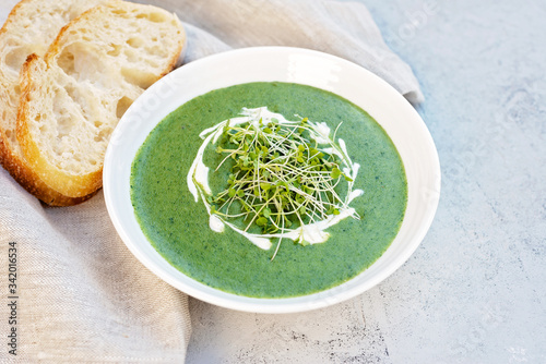 Cream soup of fresh spinach with cream and arugula microgreen in a white plate with ciabatta bread on a light gray background. Healthy and diet food.