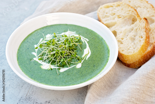 Cream soup of fresh spinach with cream and arugula microgreen in a white plate with ciabatta bread on a light gray background. Healthy and diet food.