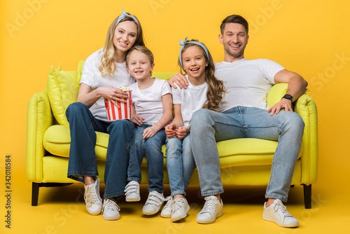 smiling parents and kids watching movie on sofa with popcorn bucket on yellow