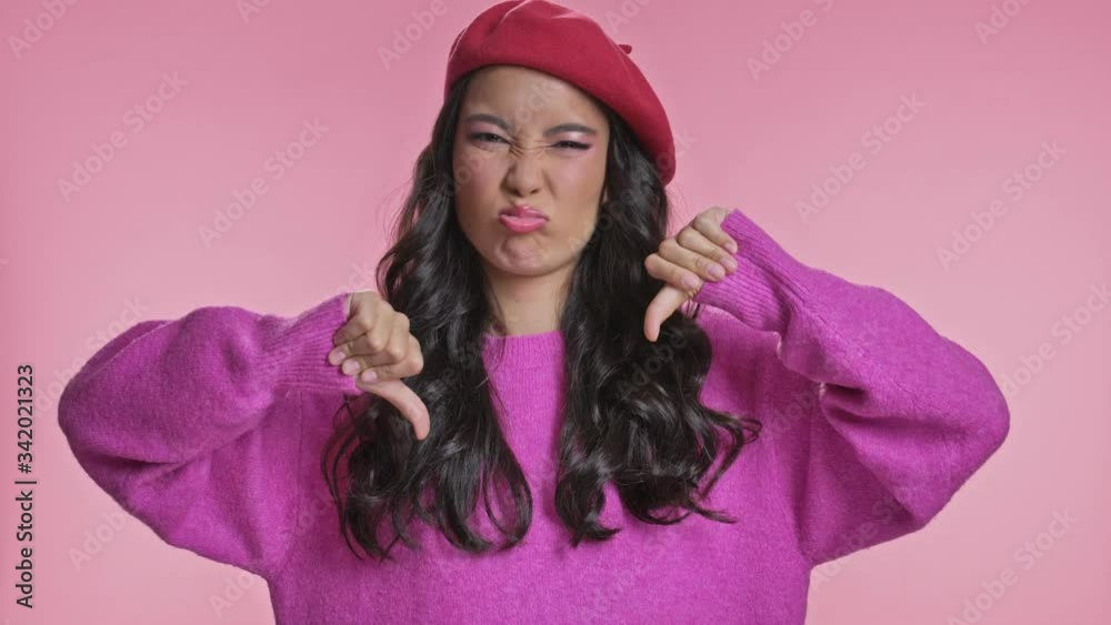 Pretty sad displeased young asian woman posing isolated over pink wall background showing thumbs down.