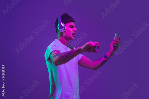 Listen to music with smartphone, dancing. Caucasian man's portrait isolated on purple studio background in pink neon light. Beautiful male model. Concept of human emotions, facial expression, sales