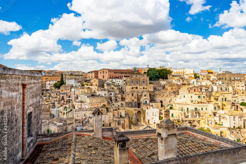 Matera old town cityscape behind a chimney with famous chimney cap, beautiful chimney decoration on a rooftop in Matera, Province of Matera, Basilicata Region, Italy under scenic cloudy summer sky