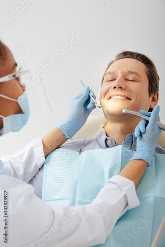 selective focus of african american dentist in medical mask and face shield holding dental instruments near happy patient