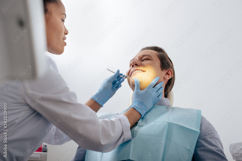 selective focus of african american dentist in latex gloves holding dental instrument near happy patient selective focus of african american dentist in latex gloves holding dental instrument near happy patient
