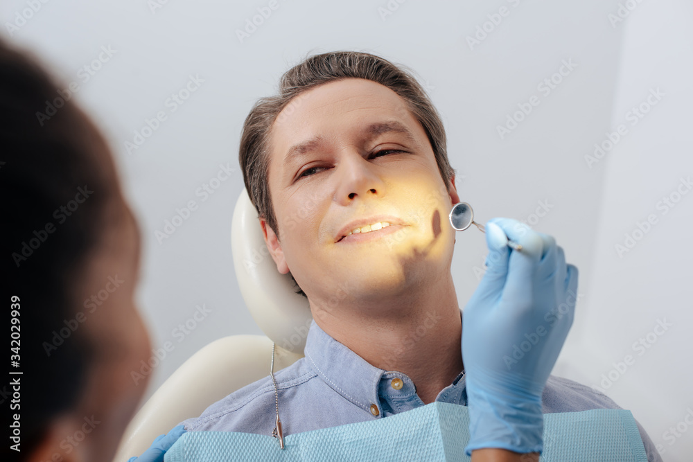 © LIGHTFIELD STUDIOS - selective focus of african american dentist in latex glove holding dental mirror near handsome patient © LIGHTFIELD STUDIOS - selective focus of african american dentist in latex glove holding dental mirror near handsome patient