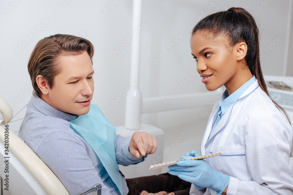 man pointing with finger at teeth palette near happy african american dentist in latex gloves