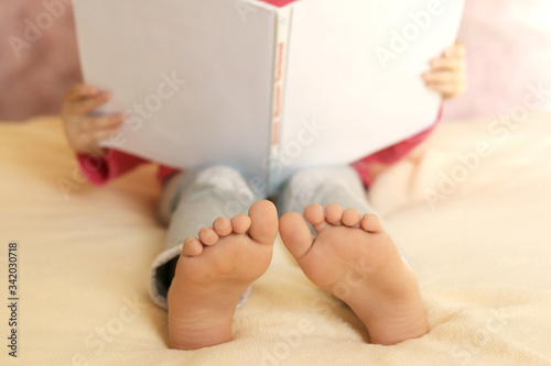 A little Caucasian girl is sitting at home on a soft bed with an open book in her hands and bare feet.Home self-isolation.Leisure activities for young children.Reading a book by a child.Selective focu